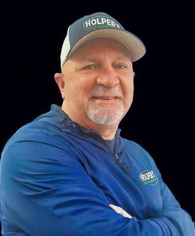 Man smiling with arms crossed wearing Holper's cap and jacket on a dark background